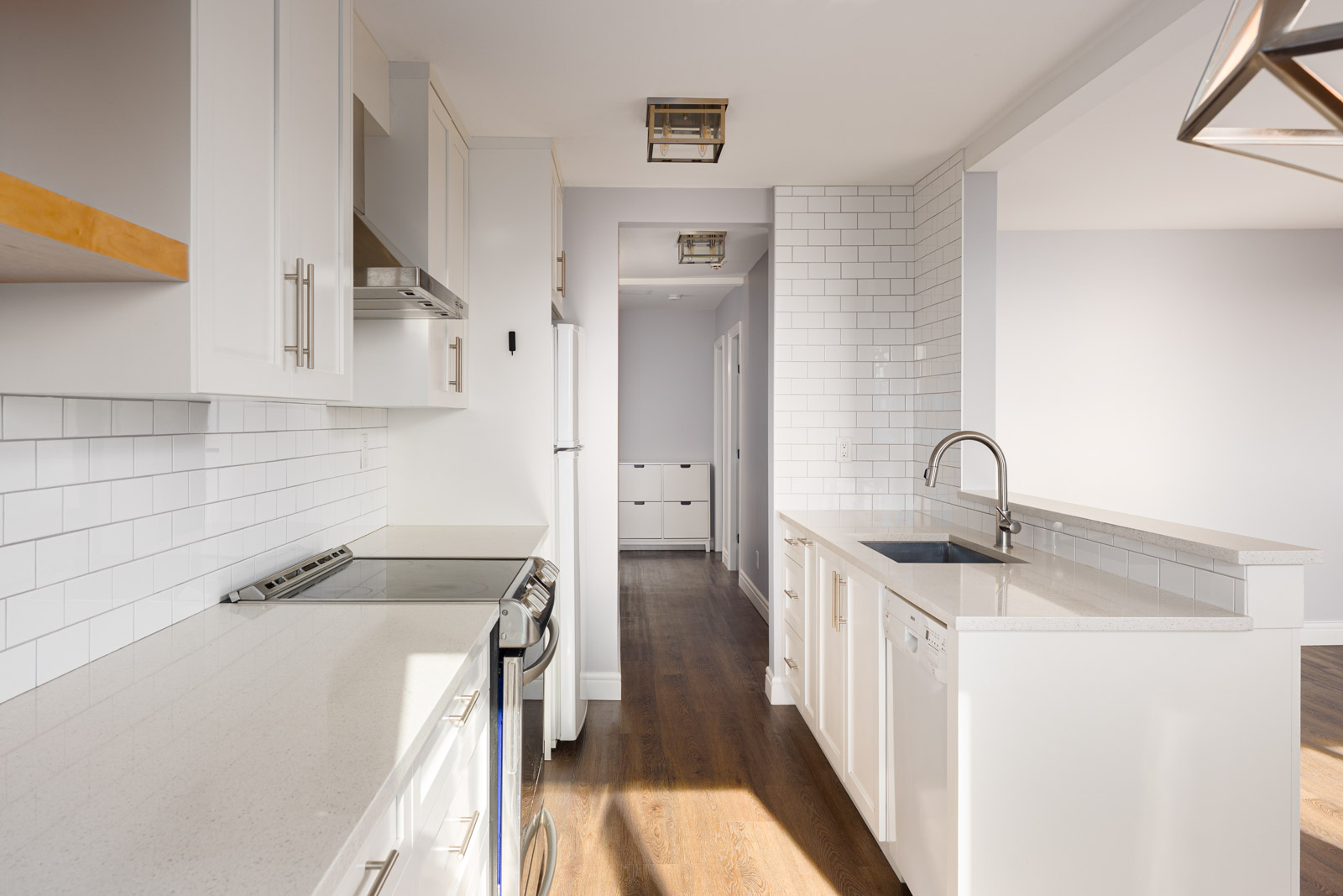 Modern kitchen with white cabinets, stainless steel appliances, subway tile backsplash, quartz countertops, and a double sink on a peninsula overlooking a hardwood floor hallway.