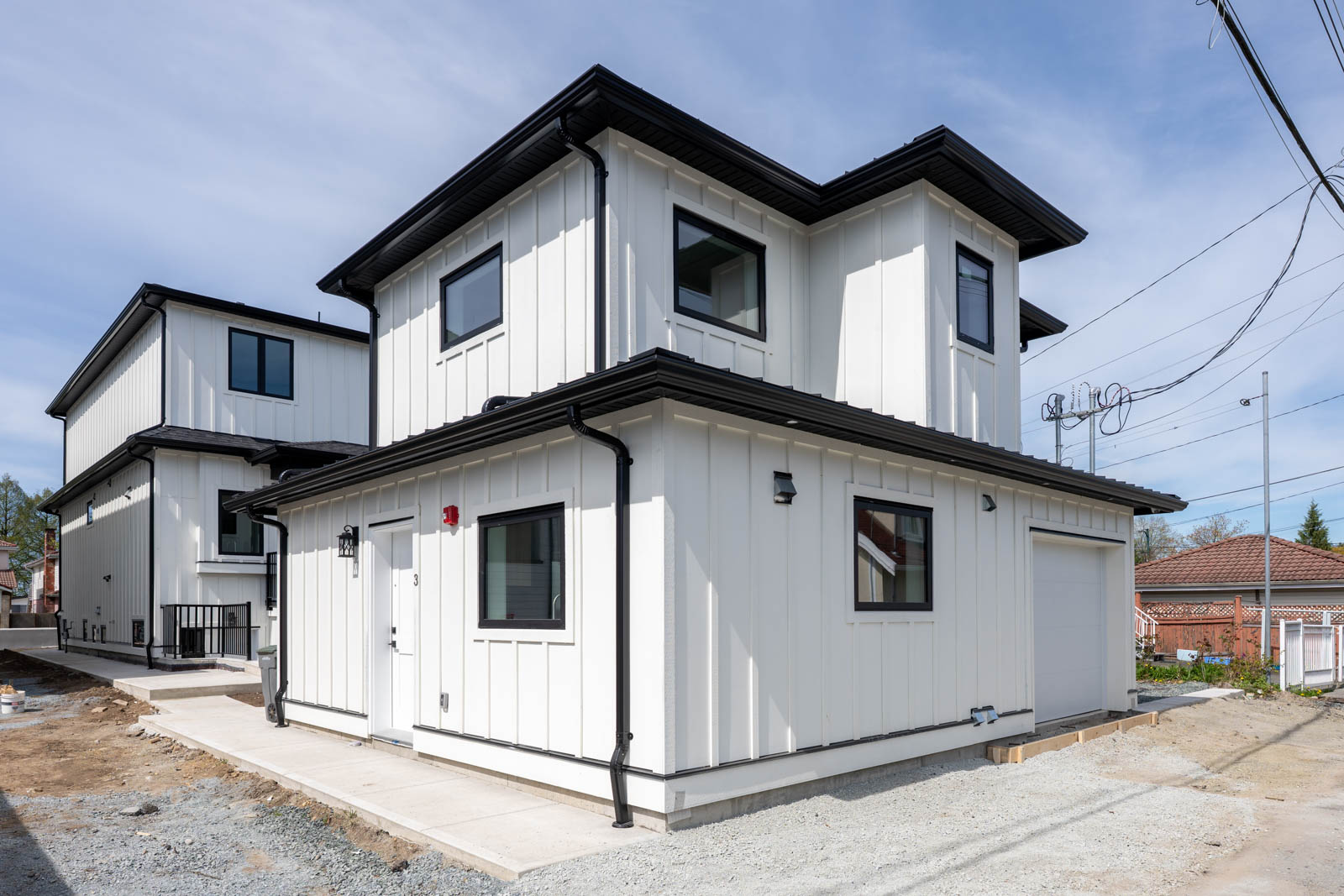 A modern two-story house with white vertical siding, black trim, and several windows, situated on a partially finished lot with a concrete walkway.