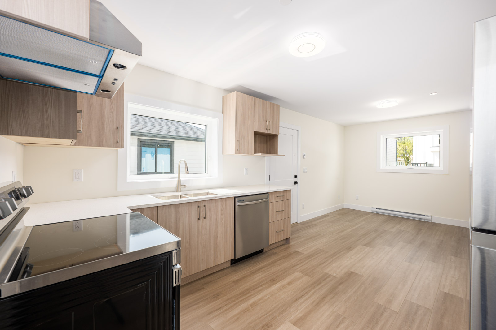 Modern kitchen with light wood cabinets, stainless steel appliances, and a window above the sink. Adjoining open space with light wood flooring and white walls.