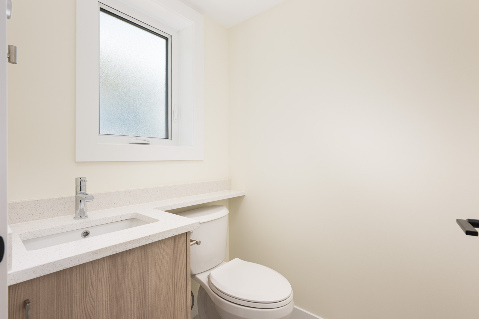 A small bathroom with a white sink, wooden vanity, and toilet under a frosted window, featuring light beige walls and a simple design.