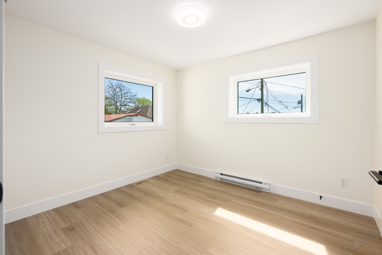 Empty room with light-colored walls, two rectangular windows, wood-look flooring, baseboard heater, and ceiling light fixture. Natural light streams in through the windows.