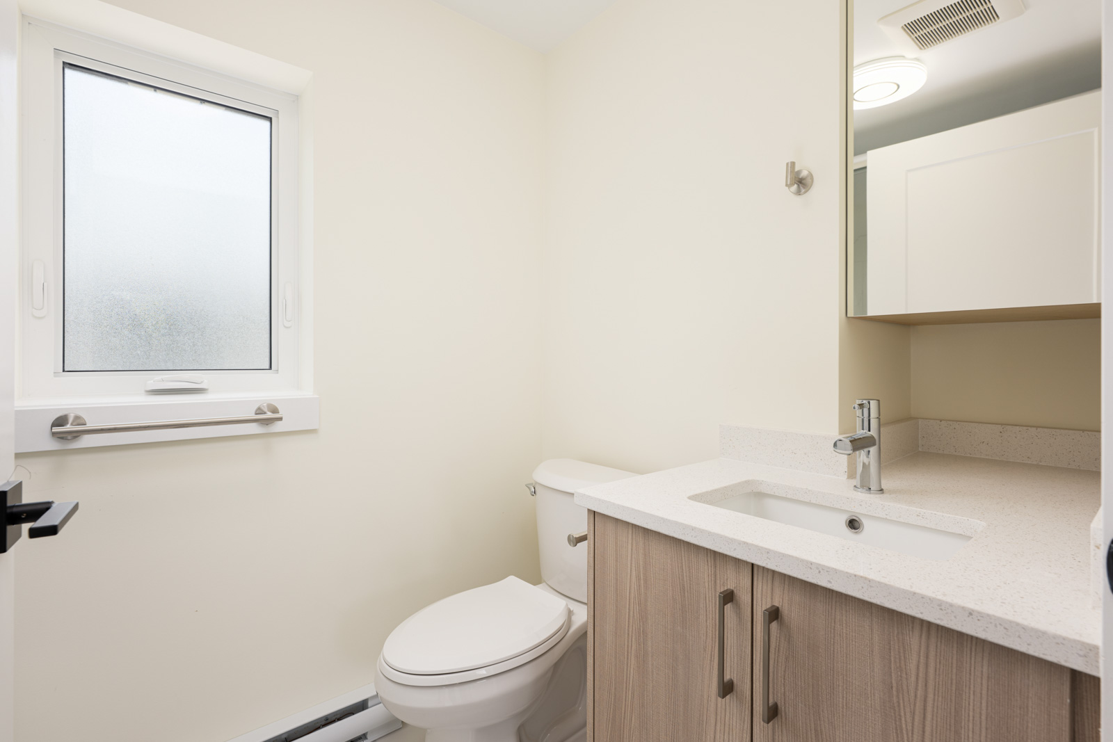 A modern bathroom with a white toilet, light wood vanity with a sink, wall mirror cabinet, frosted window, and off-white walls.
