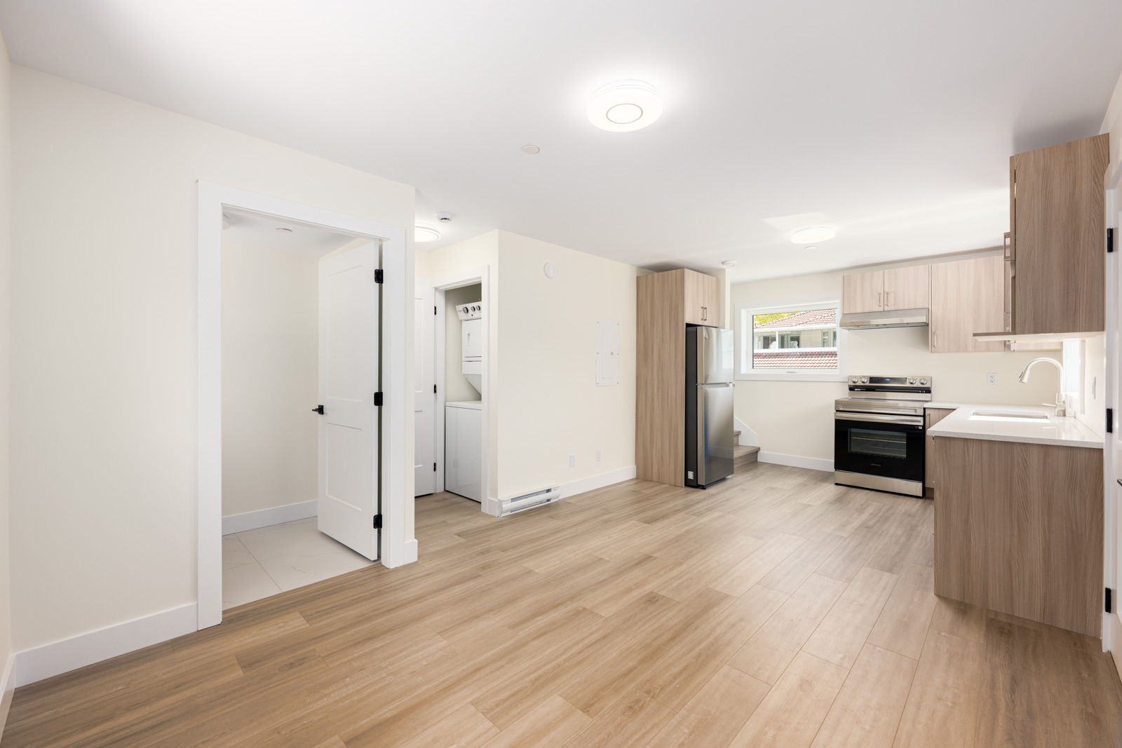 Modern kitchen and laundry area in a small apartment with light wood floors, white walls, and stainless steel appliances. Open layout with natural light from a window.