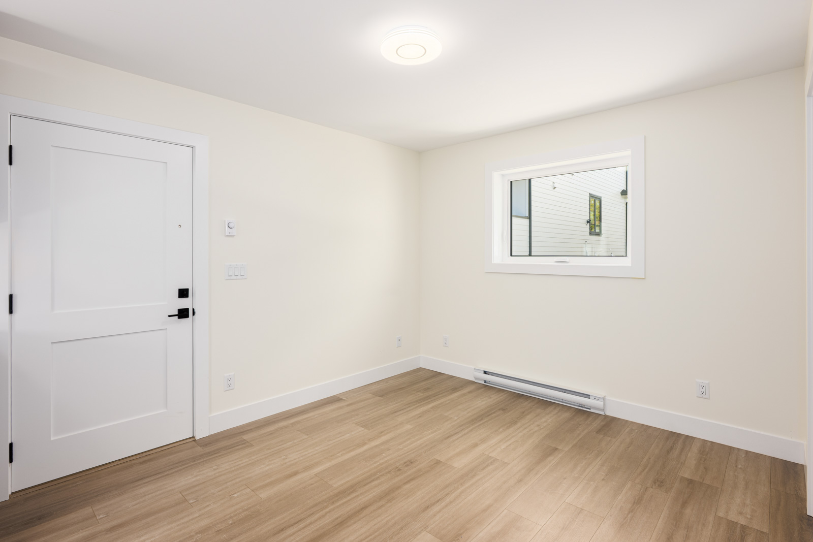 Empty room with light-colored walls, wood flooring, a white door, a window, baseboard heater, and ceiling light.