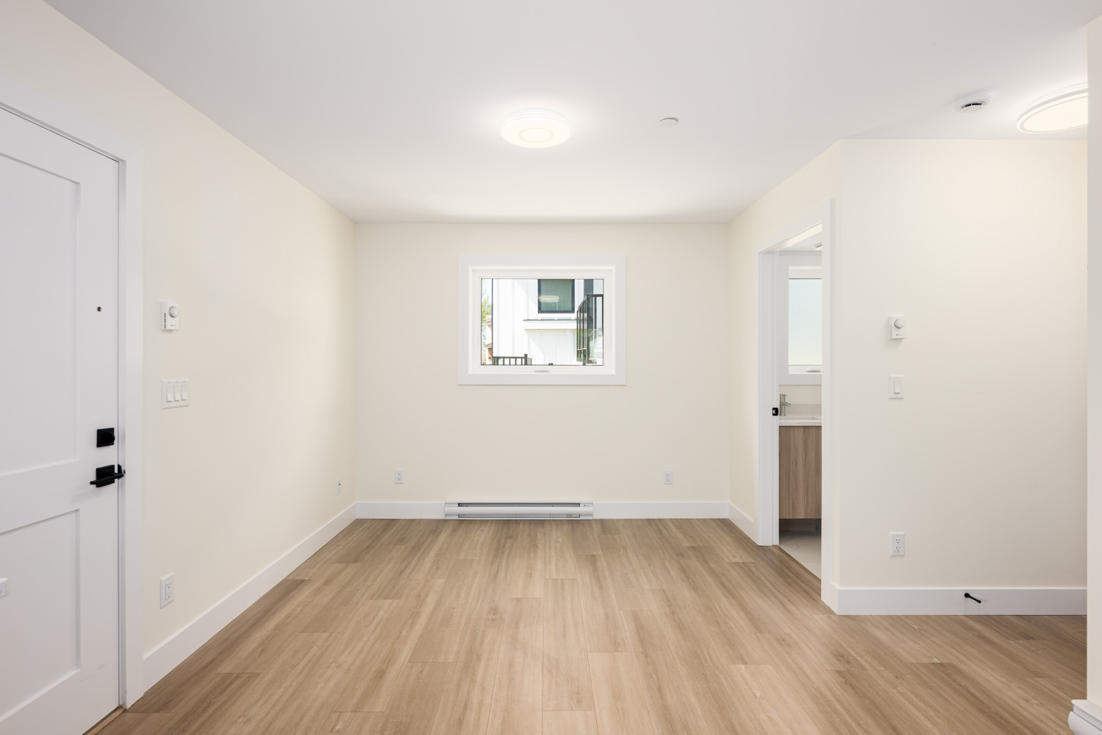 Empty room with light wood flooring, cream-colored walls, a white door on the left, a window in the center, and an open doorway to a bathroom on the right.
