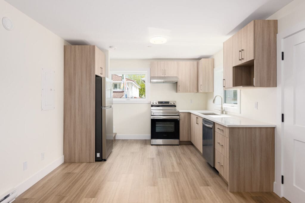 Modern kitchen with light wood cabinets, stainless steel appliances, white countertops, and light wood flooring. Large window above sink provides natural light.