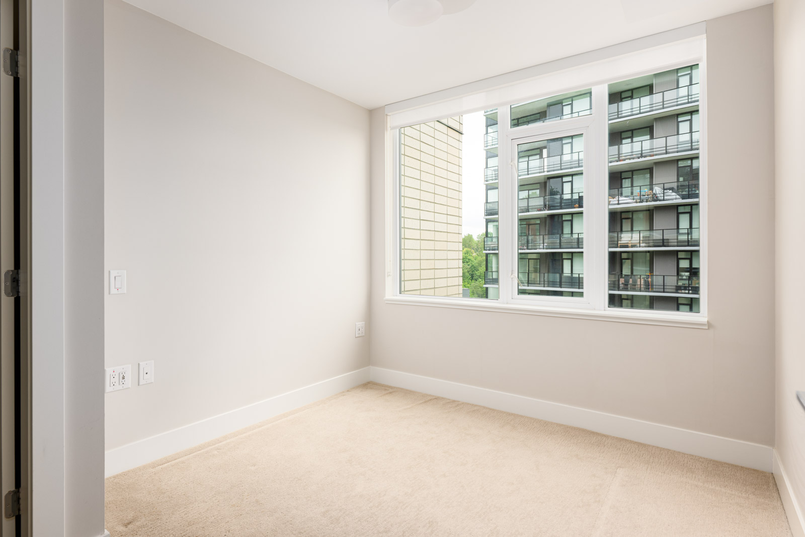 Empty, carpeted room with beige walls and a large window overlooking nearby apartment balconies and greenery.