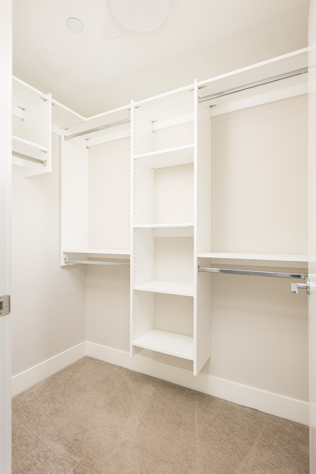 Empty walk-in closet with beige carpet, white shelves, and multiple hanging rods against light-colored walls.