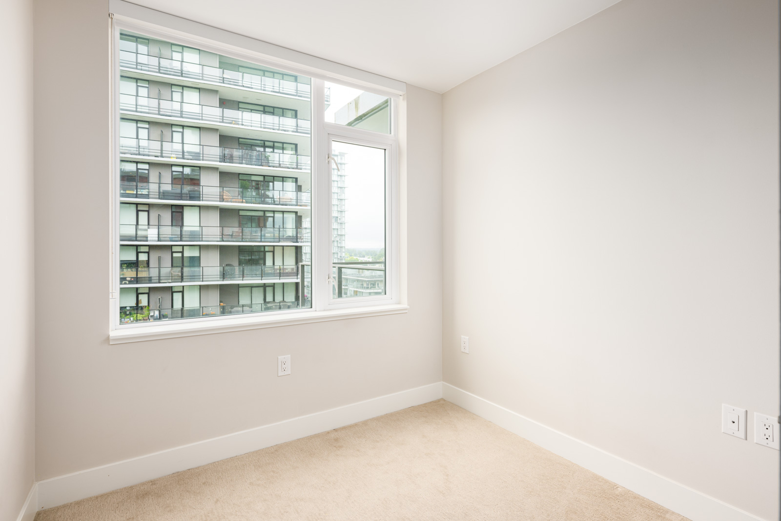 Empty room with beige carpet and white walls, featuring a large window overlooking a neighboring modern apartment building.