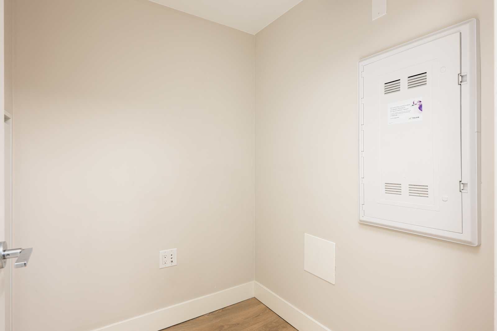 A small, empty beige corner room with a white electrical panel on the wall, an electrical outlet, and light wood flooring.