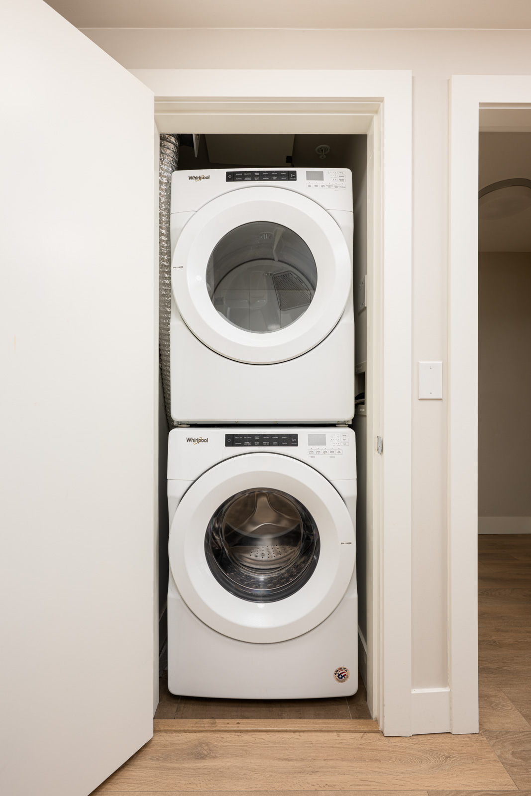 A stacked washer and dryer set in a small laundry closet with the door partially open, showing light wood flooring and white walls.