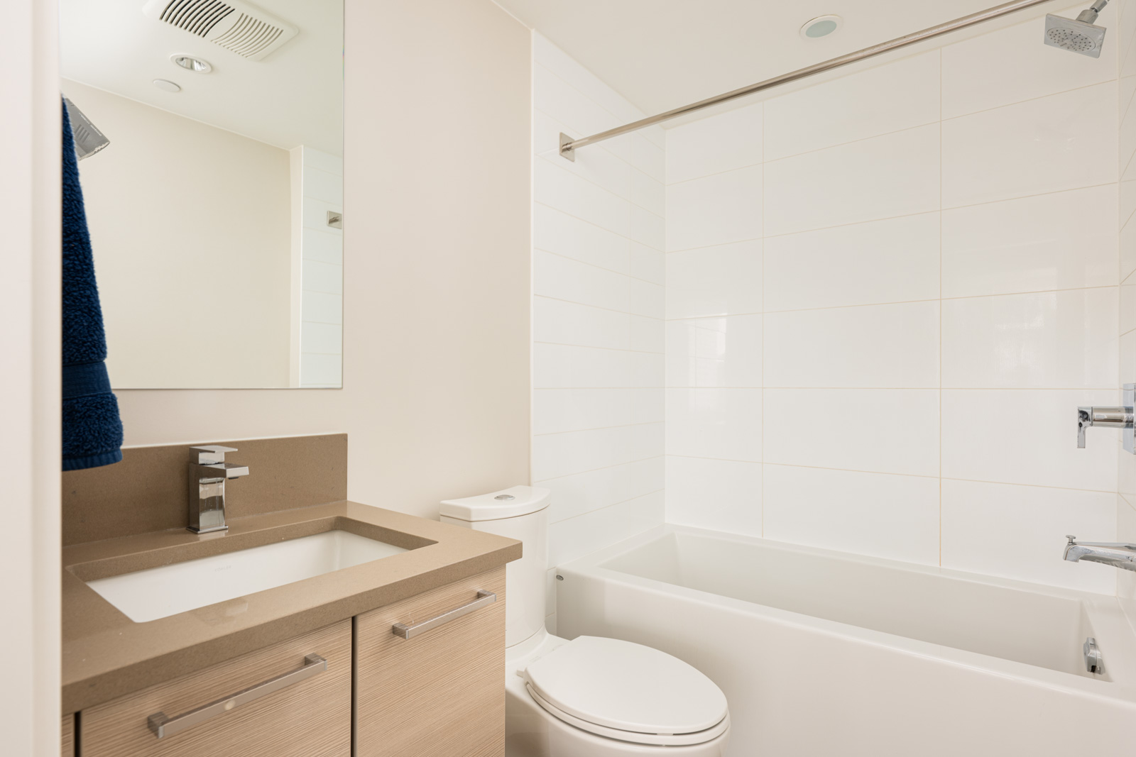 Modern bathroom with a beige vanity, rectangular sink, wall mirror, toilet, and a white bathtub with a shower curtain rod.