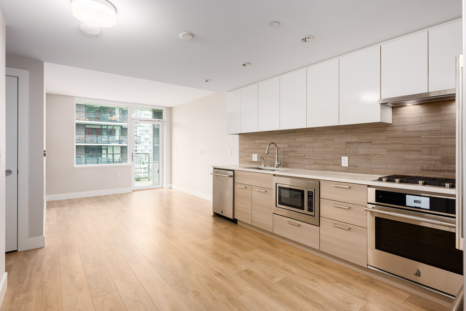 Modern apartment kitchen with light wood floors, white cabinets, built-in appliances, and large windows overlooking other buildings.
