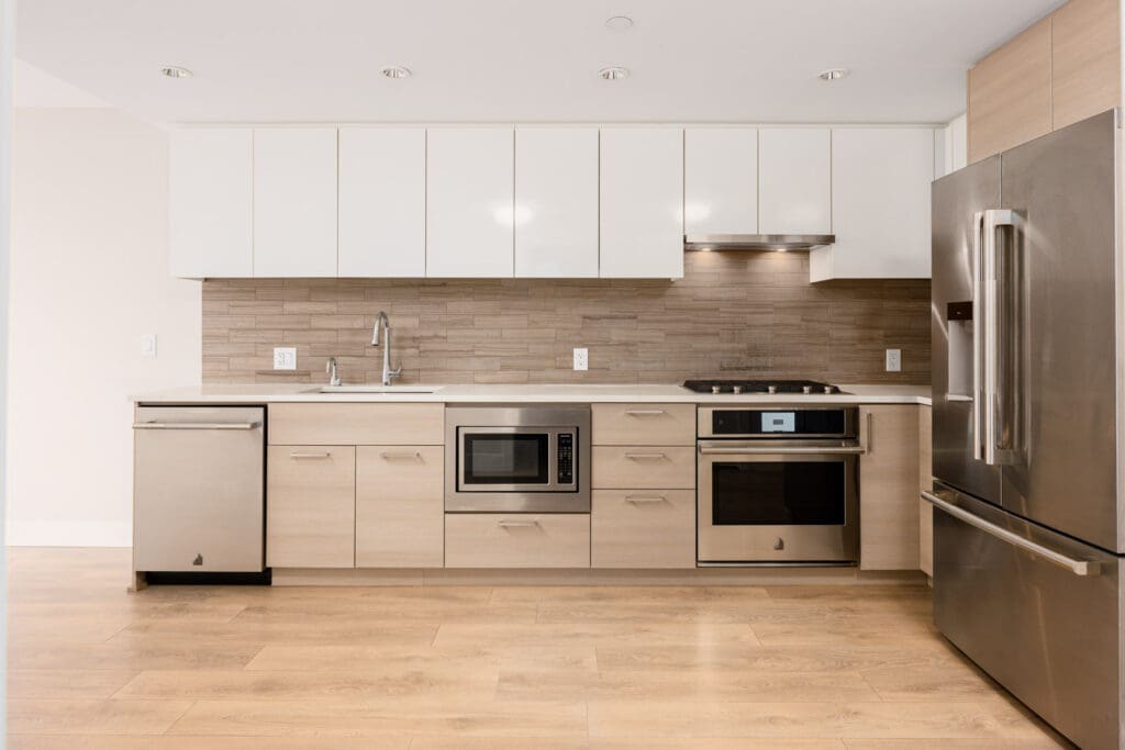 Modern kitchen with stainless steel appliances, white upper cabinets, light wood lower cabinets, and a tile backsplash, set against a hardwood floor.