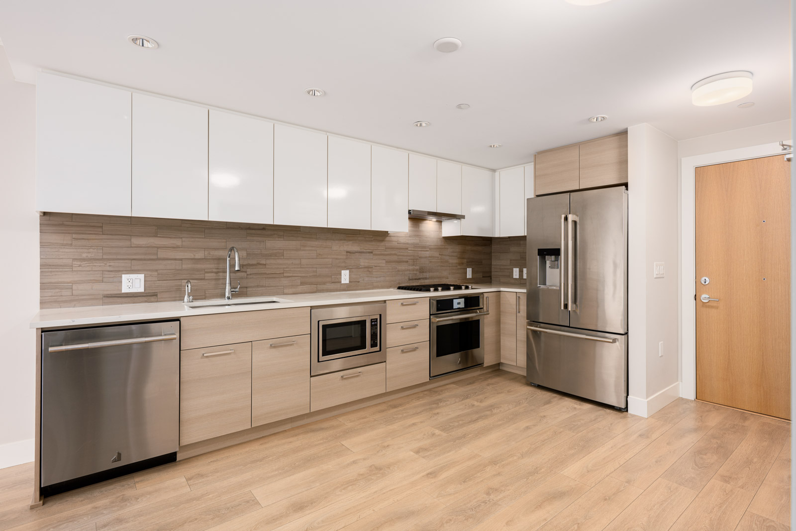 Modern kitchen with light wood floors, white and wood cabinetry, stainless steel appliances, and a tile backsplash.