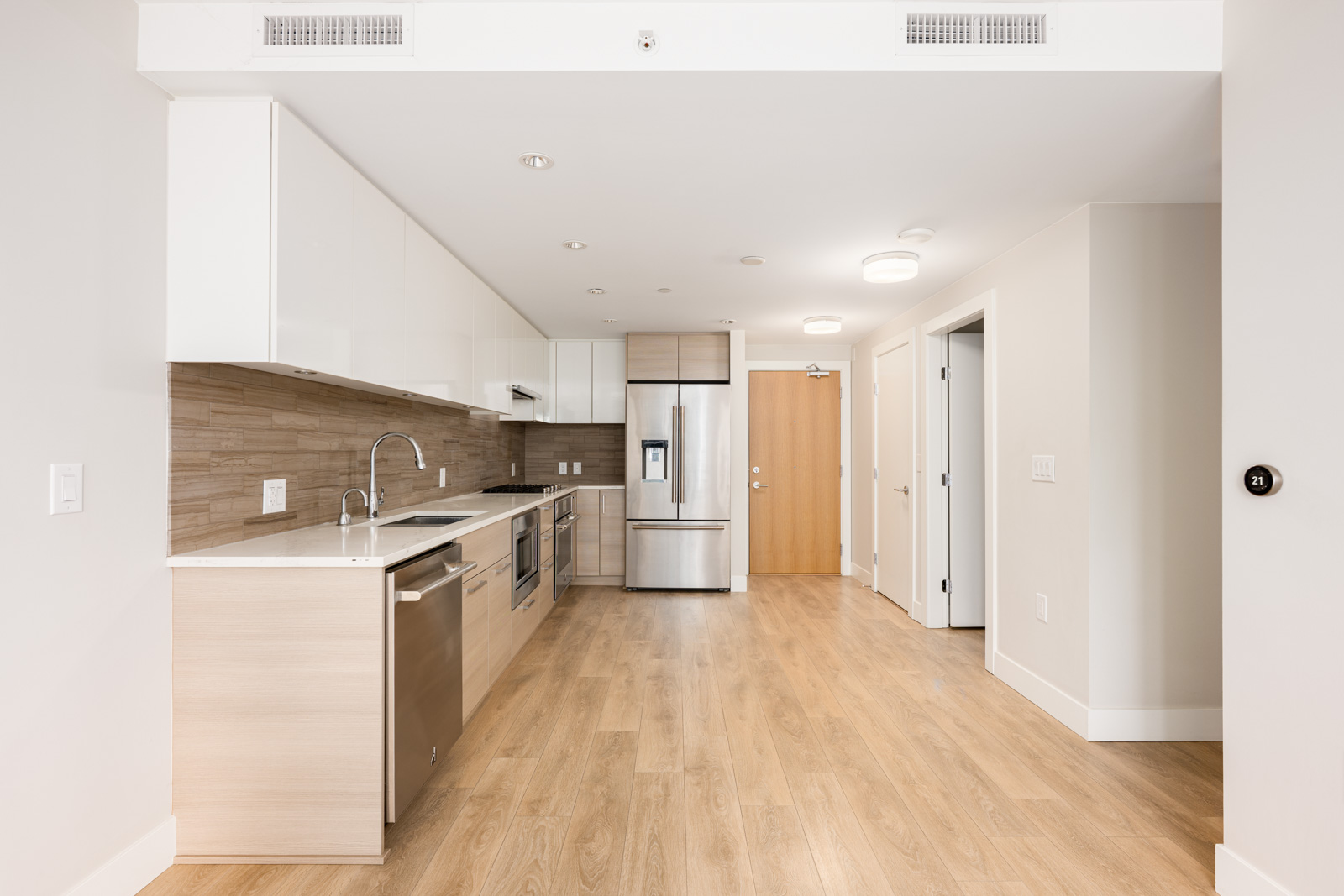 Modern kitchen with light wood floors, white cabinets, stainless steel appliances, and a wood-textured backsplash, adjacent to a hallway with doors.