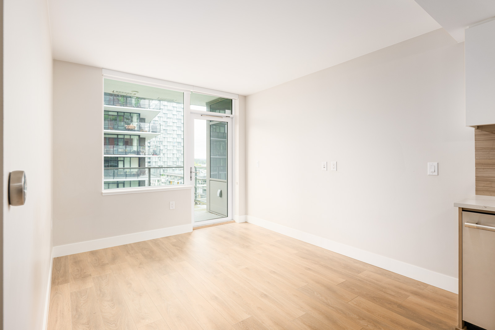 Unfurnished, modern apartment room with light wood floors, large window and glass door leading to a balcony, overlooking a contemporary building exterior.