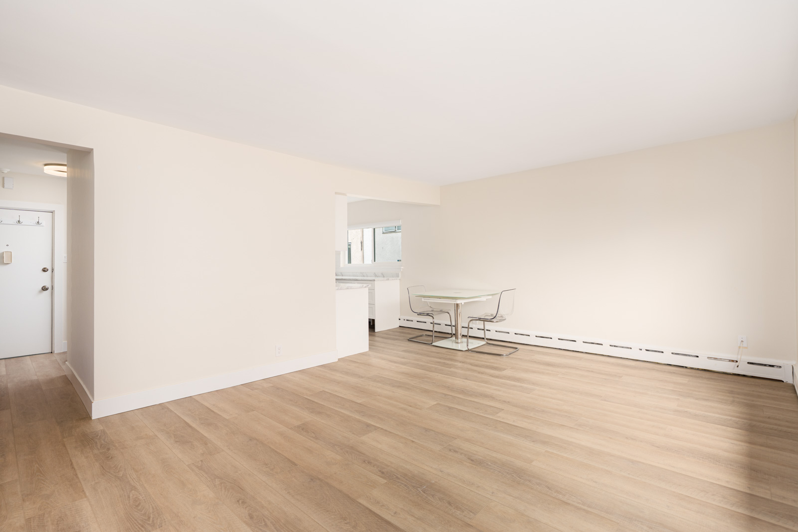 Empty room with light wood flooring, white walls, and an open doorway leading to a kitchen. A small table with two chairs is placed near the back wall.