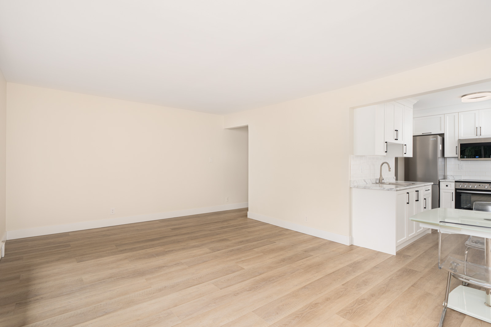 Empty room with light wood flooring and cream walls, adjacent to a white kitchen featuring stainless steel appliances and a glass table with chairs.