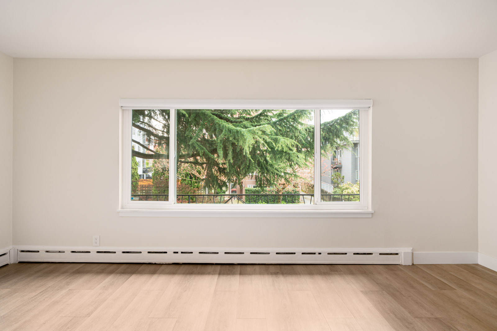 Empty room with light wood flooring and a large window revealing a view of greenery outside. Beige walls and baseboard heater visible.