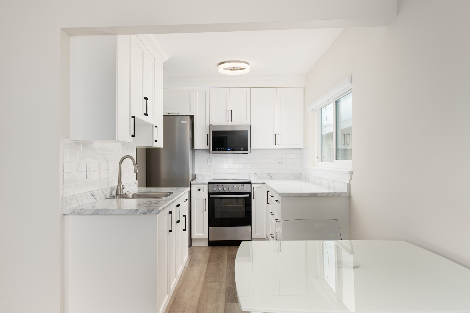Modern kitchen with white cabinets, stainless steel appliances, marble countertops, and a glass-top table. Wood flooring complements the neutral color scheme.