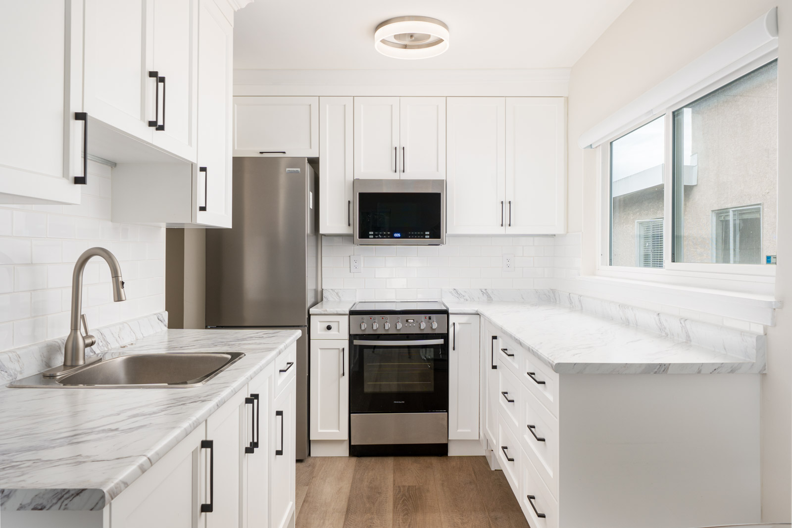 Modern kitchen with white cabinets, stainless steel appliances, a marble countertop, and wooden flooring. A window on the right provides natural light.