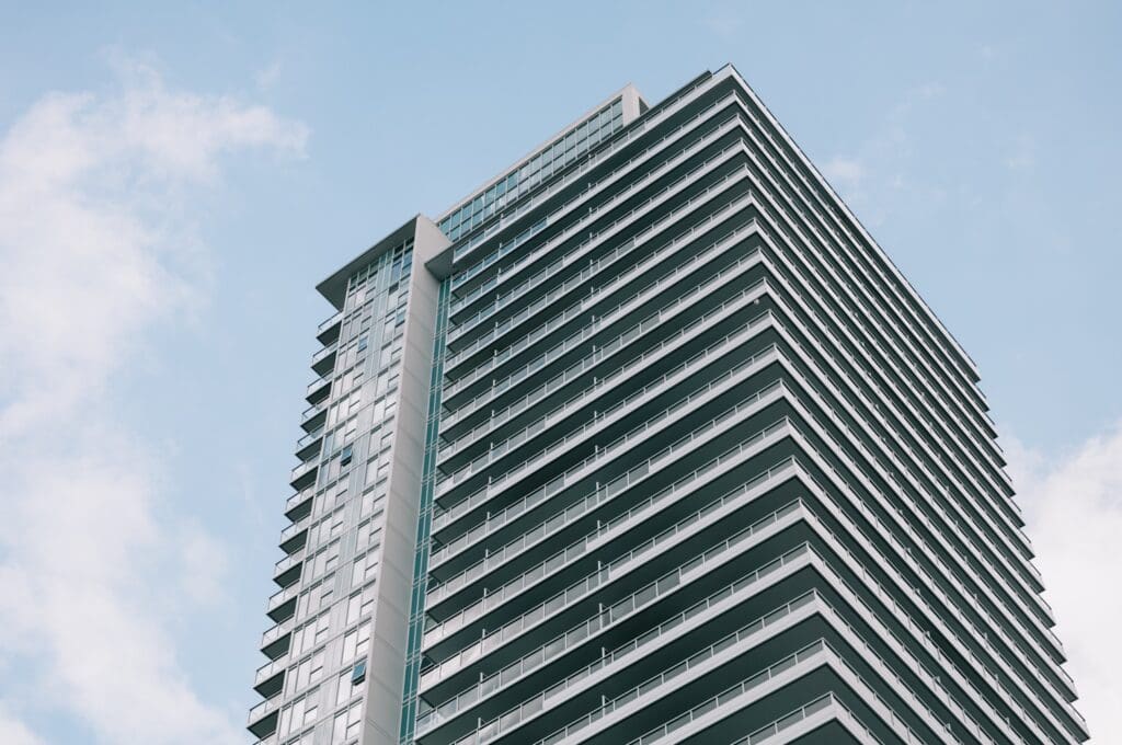 A modern high-rise building with numerous balconies and glass windows against a blue sky with some clouds.