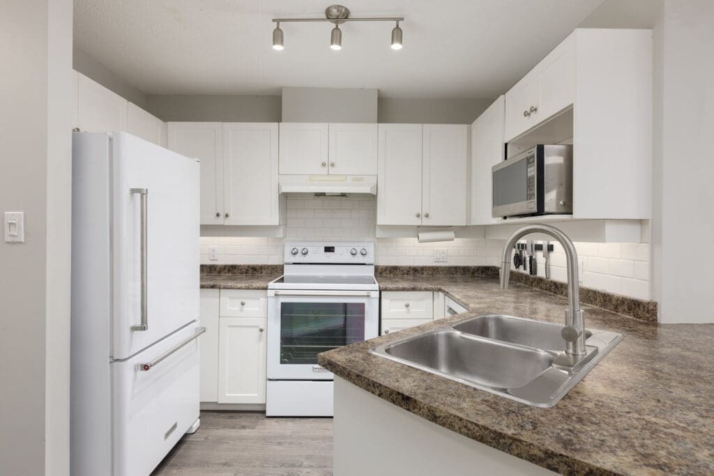 Modern kitchen with white cabinets, a white refrigerator, stove, microwave, and a dual-basin sink on a brown marble countertop under track lighting.