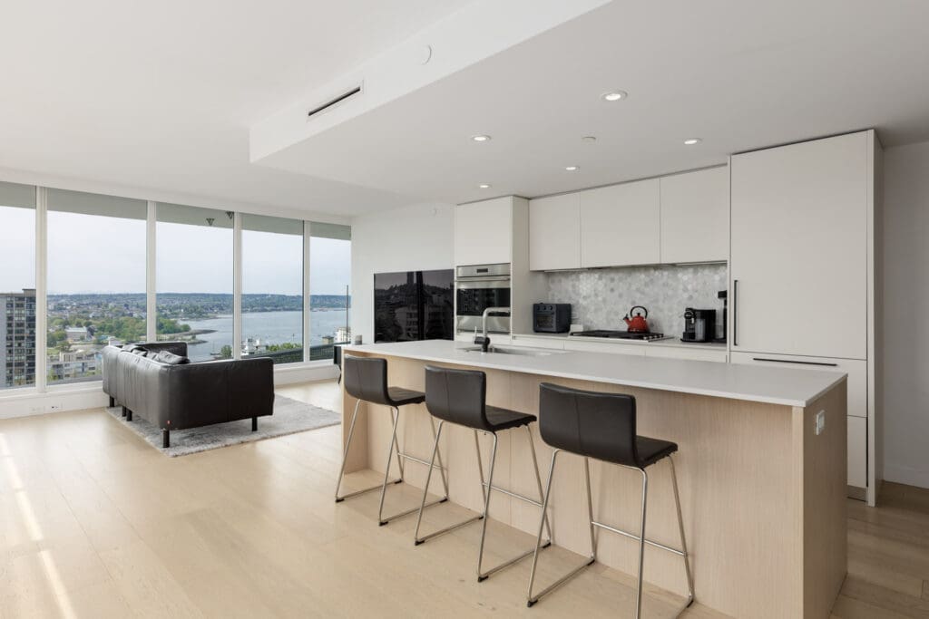 Modern open-concept kitchen with white cabinets, island with three bar stools, and living area; large windows offer a view of water and cityscape.