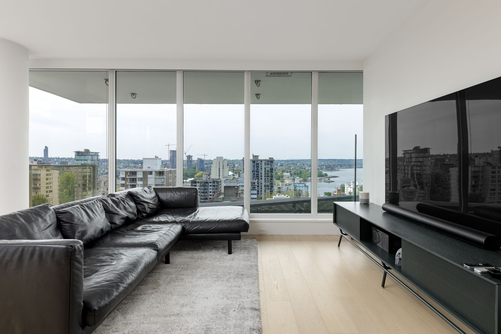 Modern living room with a black leather sectional sofa, large windows overlooking a cityscape and water, a grey rug, and a TV on a low media console.