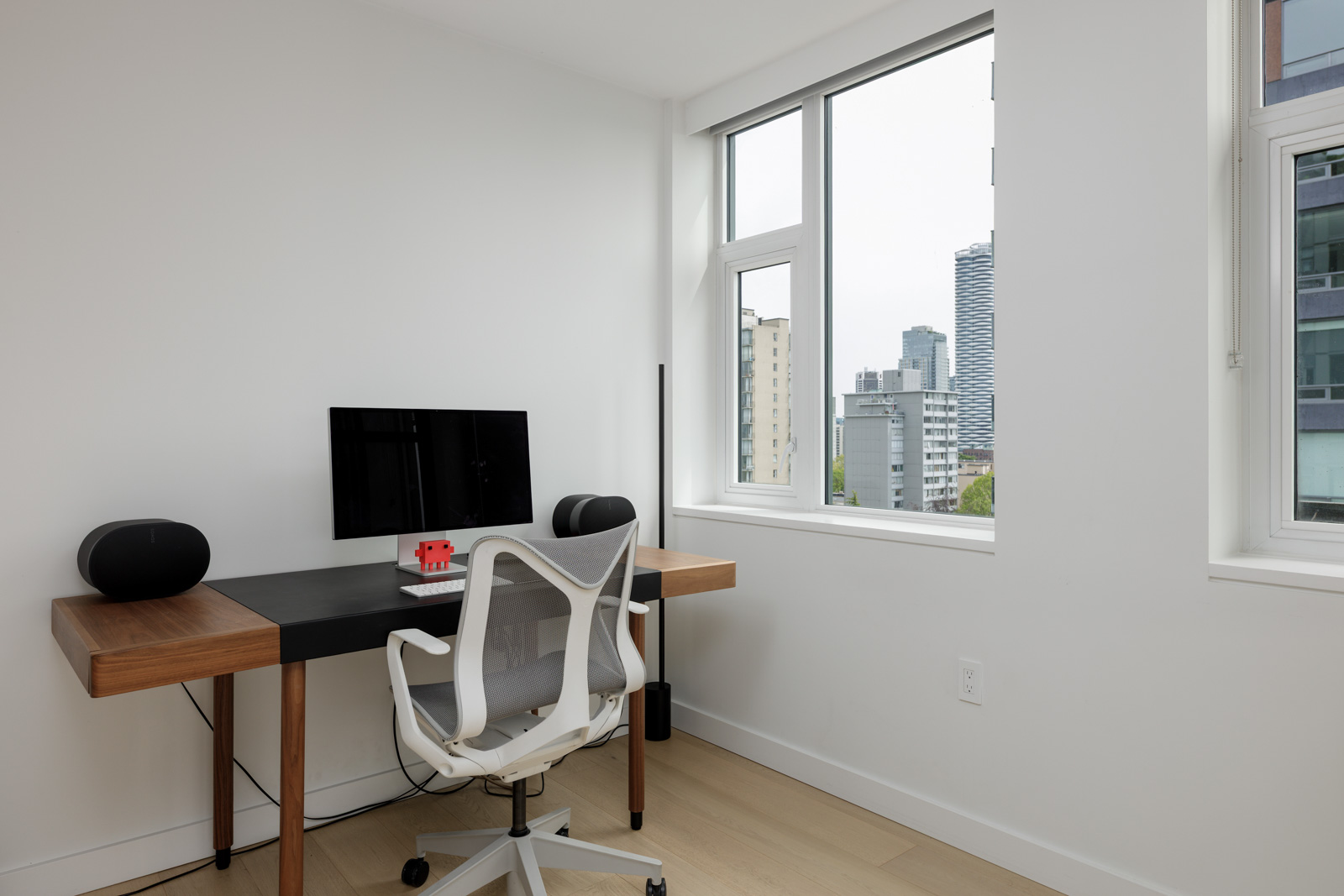 A minimalist home office with a wooden desk, computer monitor, chair, two speakers, and a window with a city view.