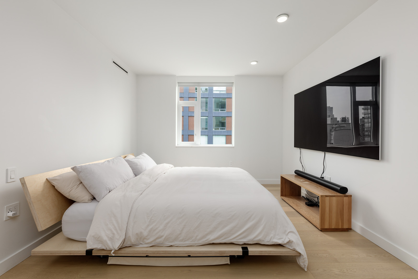 Minimalist bedroom with a low wooden bed, white bedding, a large wall-mounted TV, and a small wooden TV stand. A window lets in natural light and shows a view of nearby buildings.