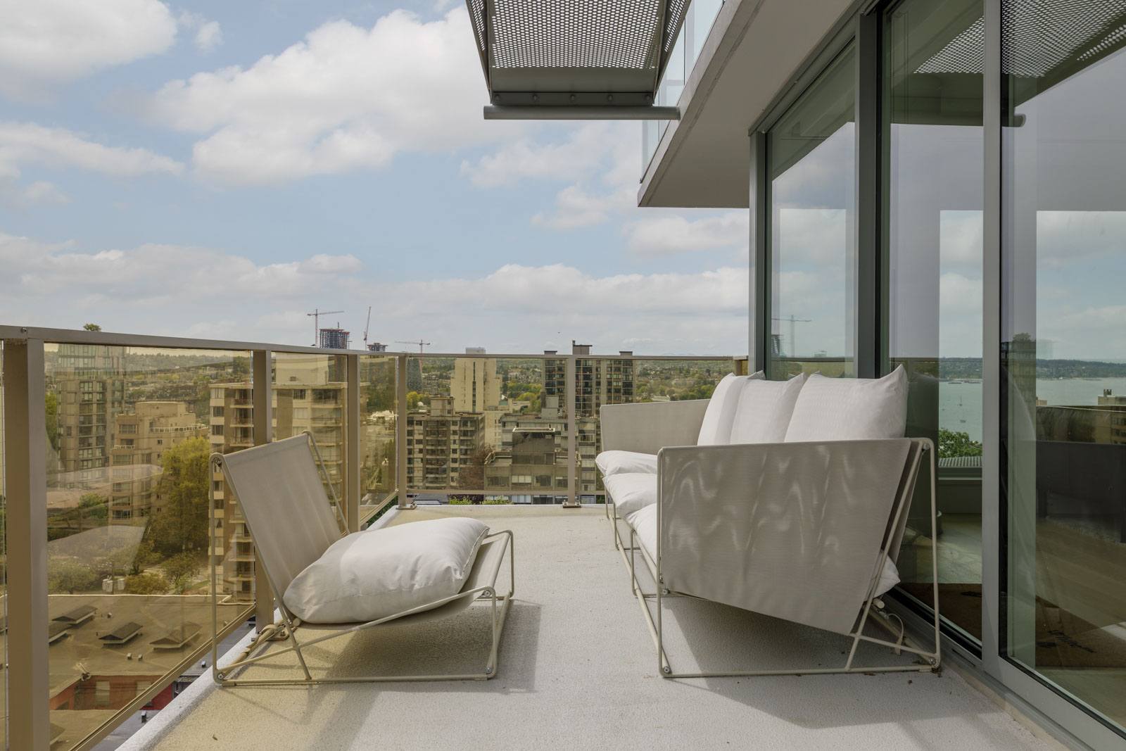 Modern balcony with glass railing, two white lounge chairs, and a glass door, overlooking city buildings under a partly cloudy sky.