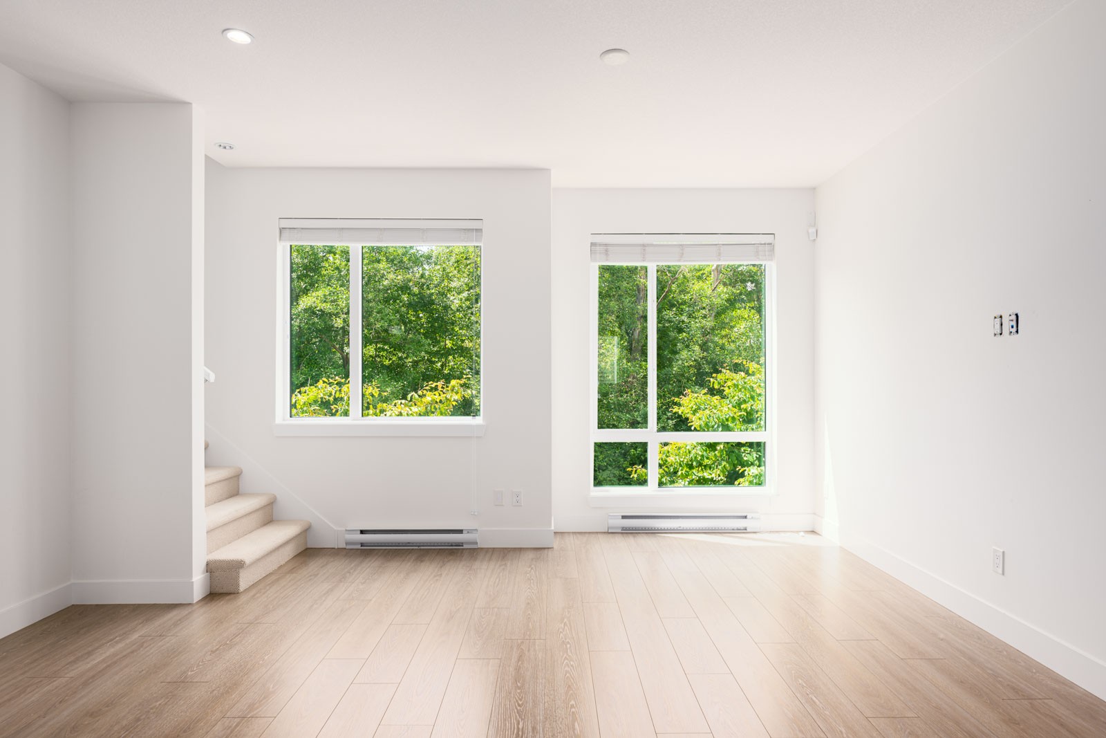 An empty, sunlit room with light wood flooring, two large windows showing green trees outside, and a staircase on the left.