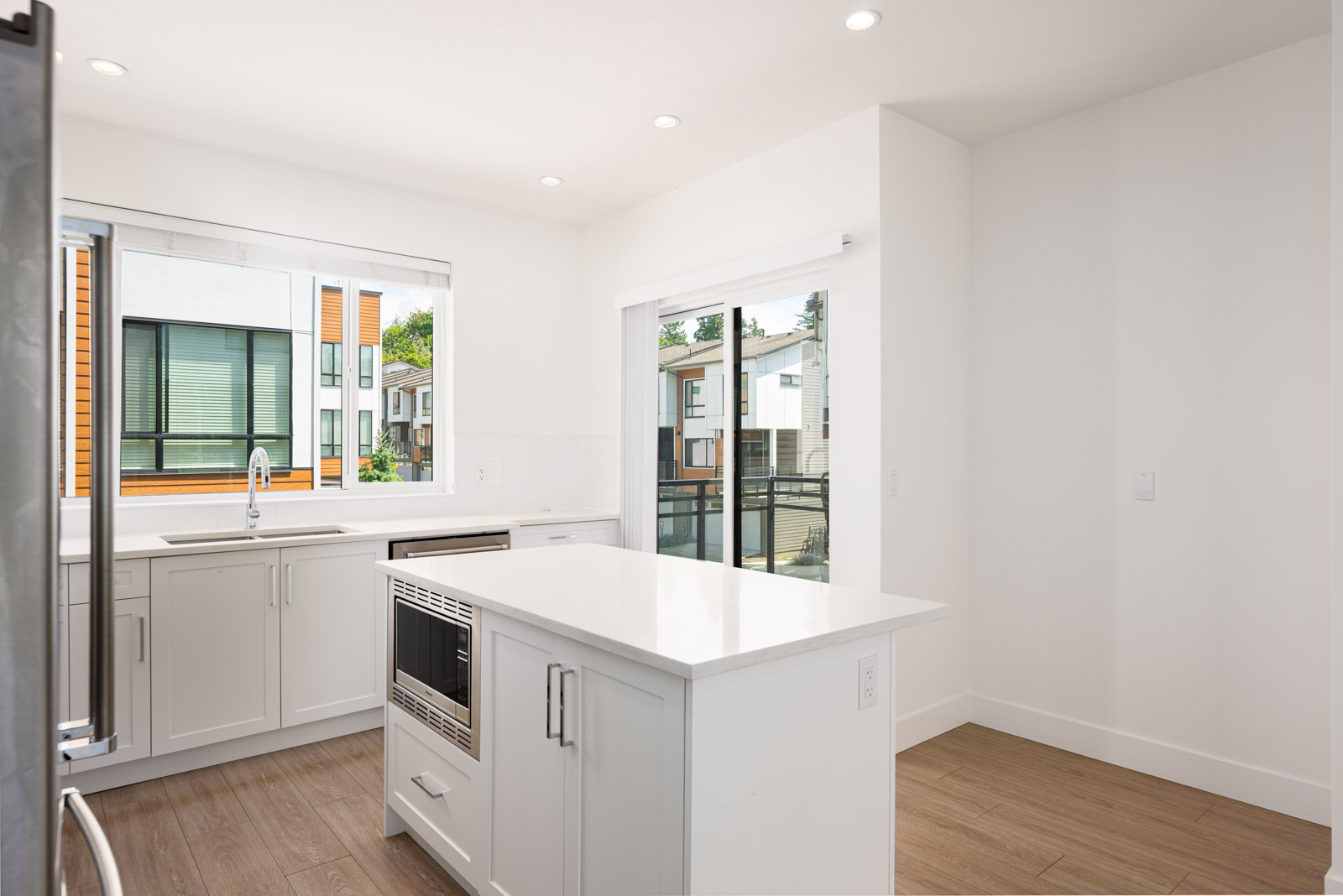 Modern kitchen with white cabinets, center island, stainless steel appliances, large window, and sliding glass door leading to a balcony.
