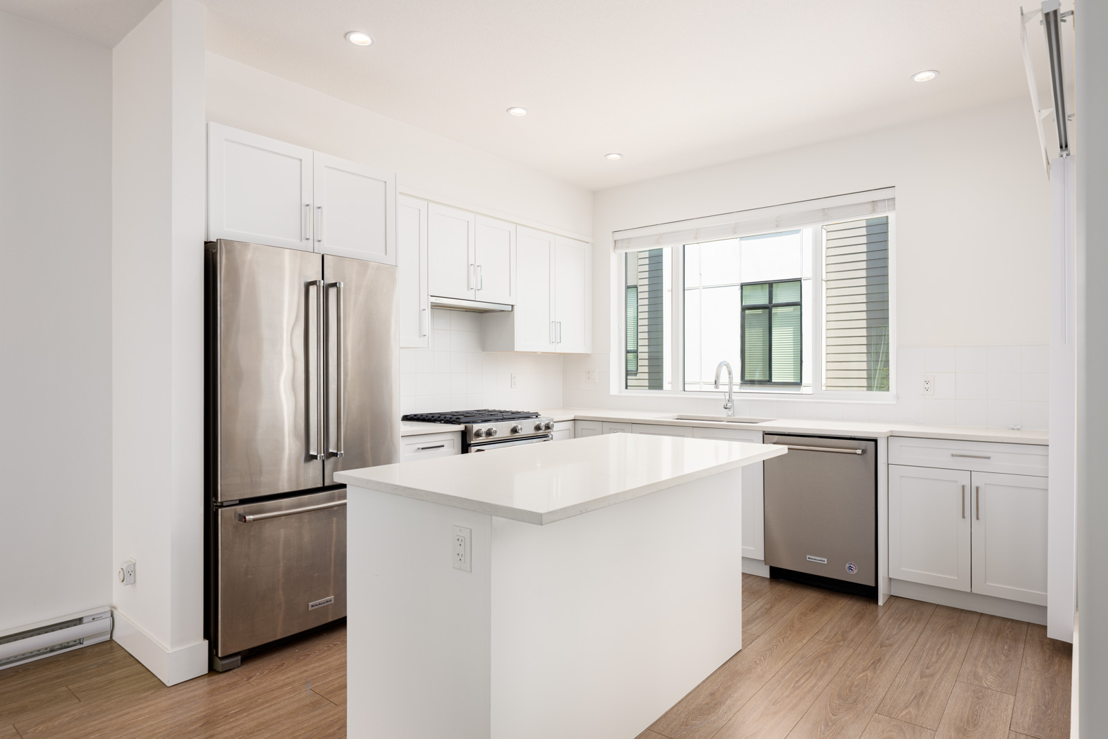 Modern kitchen with white cabinets, stainless steel appliances, an island with a quartz countertop, a large window, and light wood flooring.