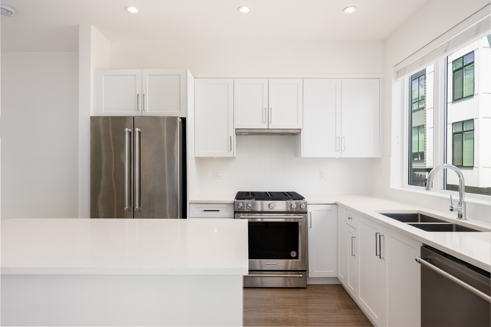 Modern kitchen with white cabinets, stainless steel refrigerator and stove, quartz countertops, large window, and a double sink with a chrome faucet.