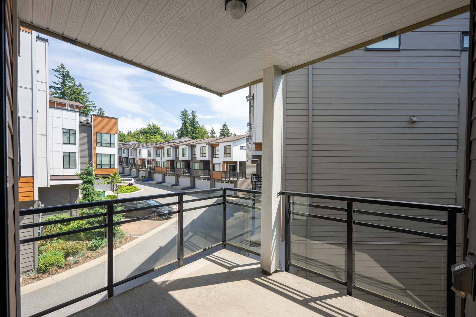 View from a covered balcony with glass and metal railing, overlooking a residential street lined with modern townhouses and trees.