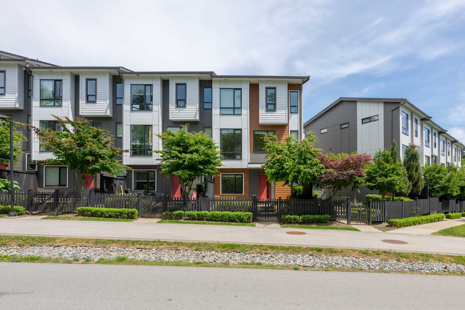 Modern three-story townhouse buildings with large windows, front yards, and black picket fences along a sidewalk under a partly cloudy sky.