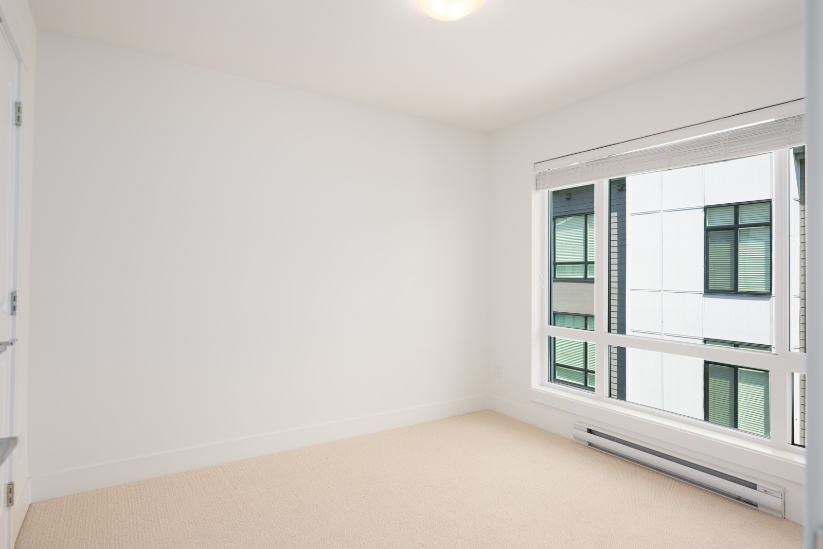 Empty room with white walls, beige carpet, a large window with blinds, and a view of an adjacent building with green-trimmed windows.