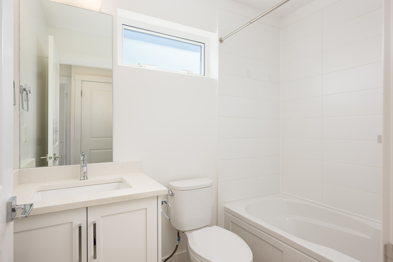 A modern white bathroom with a sink, toilet, and bathtub; a large mirror and a small horizontal window above the toilet let in natural light.