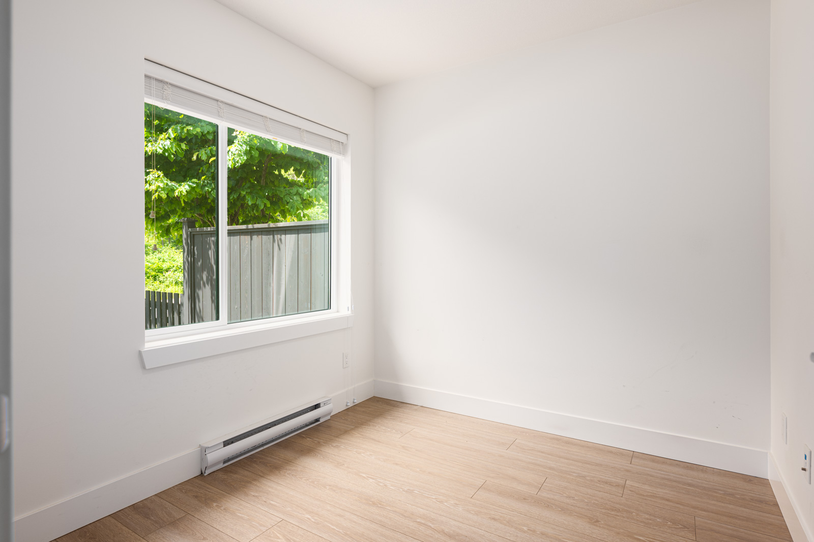 Empty room with light-colored walls and wood-style flooring, a large window with a view of greenery, and a baseboard heater beneath the window.