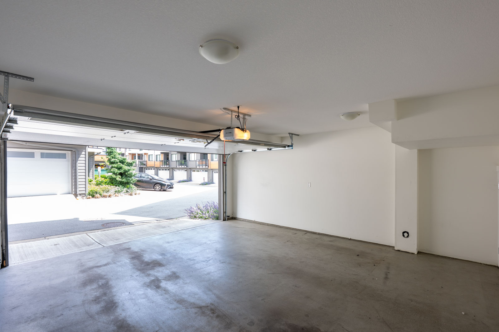 Empty two-car garage with gray concrete floor, white walls, ceiling lights, and an open garage door showing a driveway and neighboring buildings.
