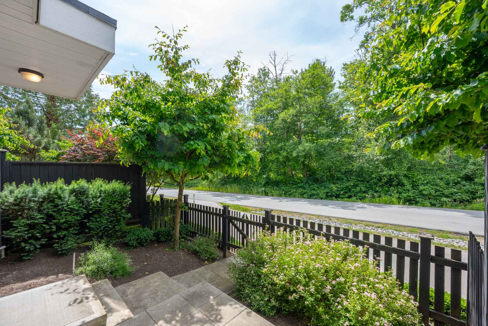View from a front porch showing concrete steps, a black picket fence, green bushes and trees, with a road and more greenery in the background.