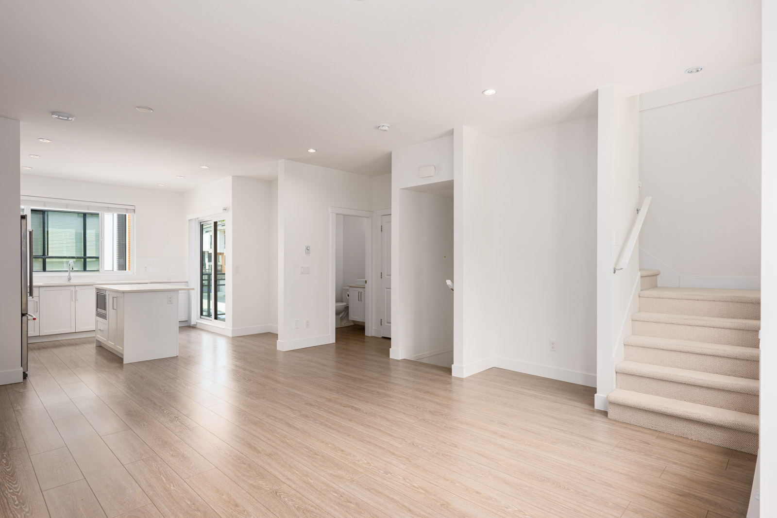 Bright, empty modern living space with light wood floors, white walls, kitchen area, sliding glass doors, and a carpeted staircase.