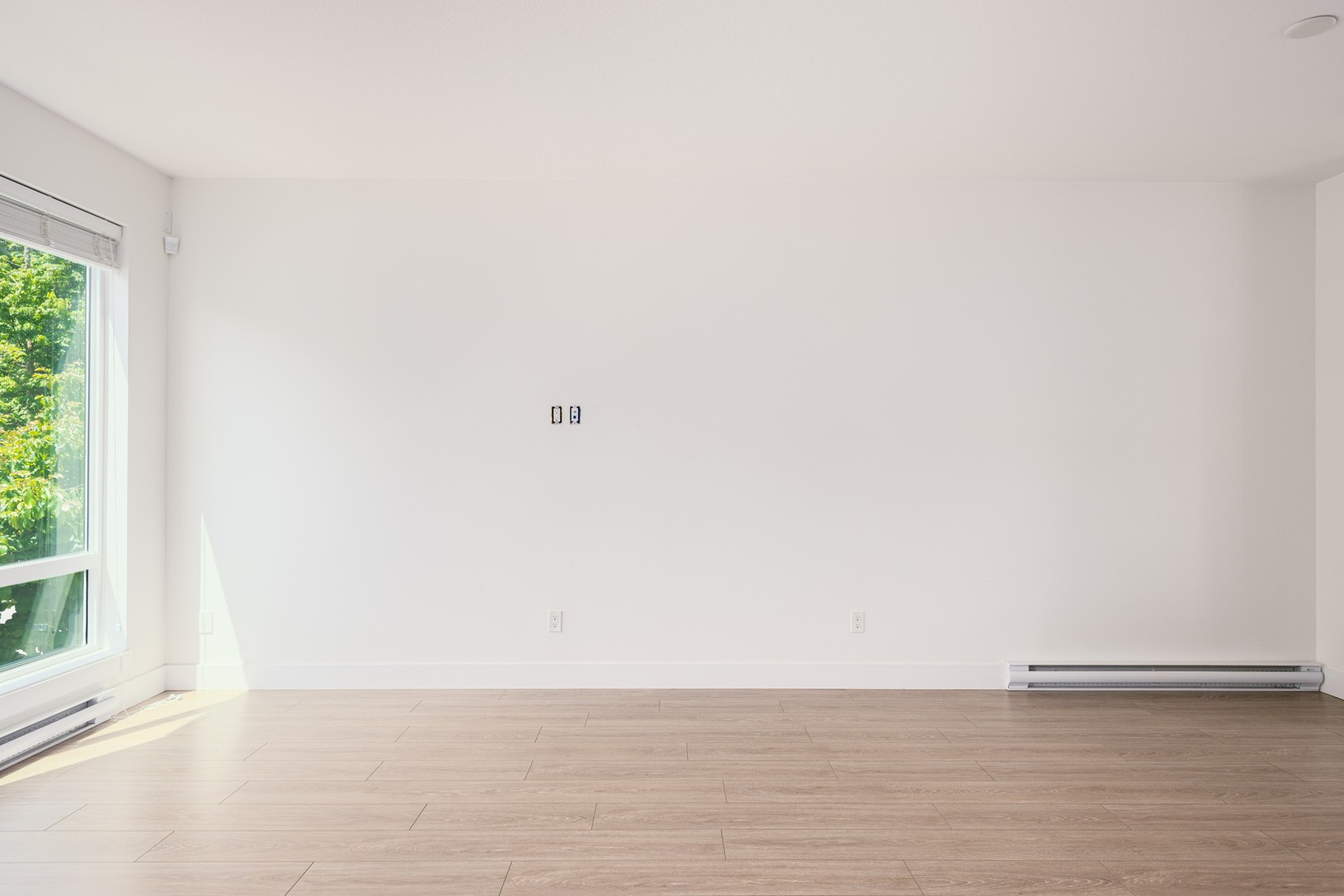 An empty room with white walls, light wood flooring, a large window on the left, and baseboard heating along the right wall.