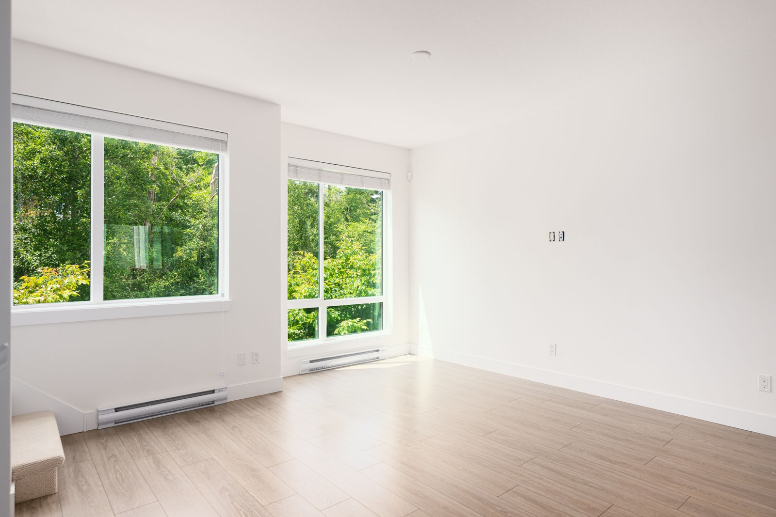 Empty, unfurnished room with large windows, white walls, and light wood floors. Green trees are visible outside the windows, and natural light fills the space.