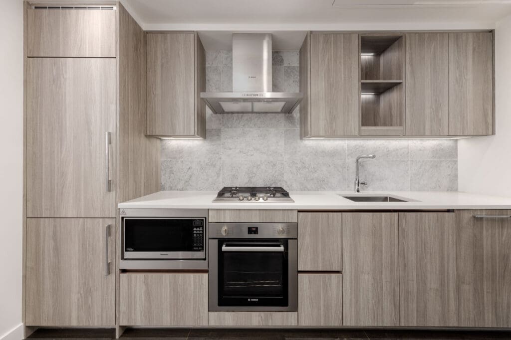 Modern kitchen with light wood cabinets, built-in appliances including oven, microwave, gas cooktop, and range hood, white countertop, and marble backsplash.