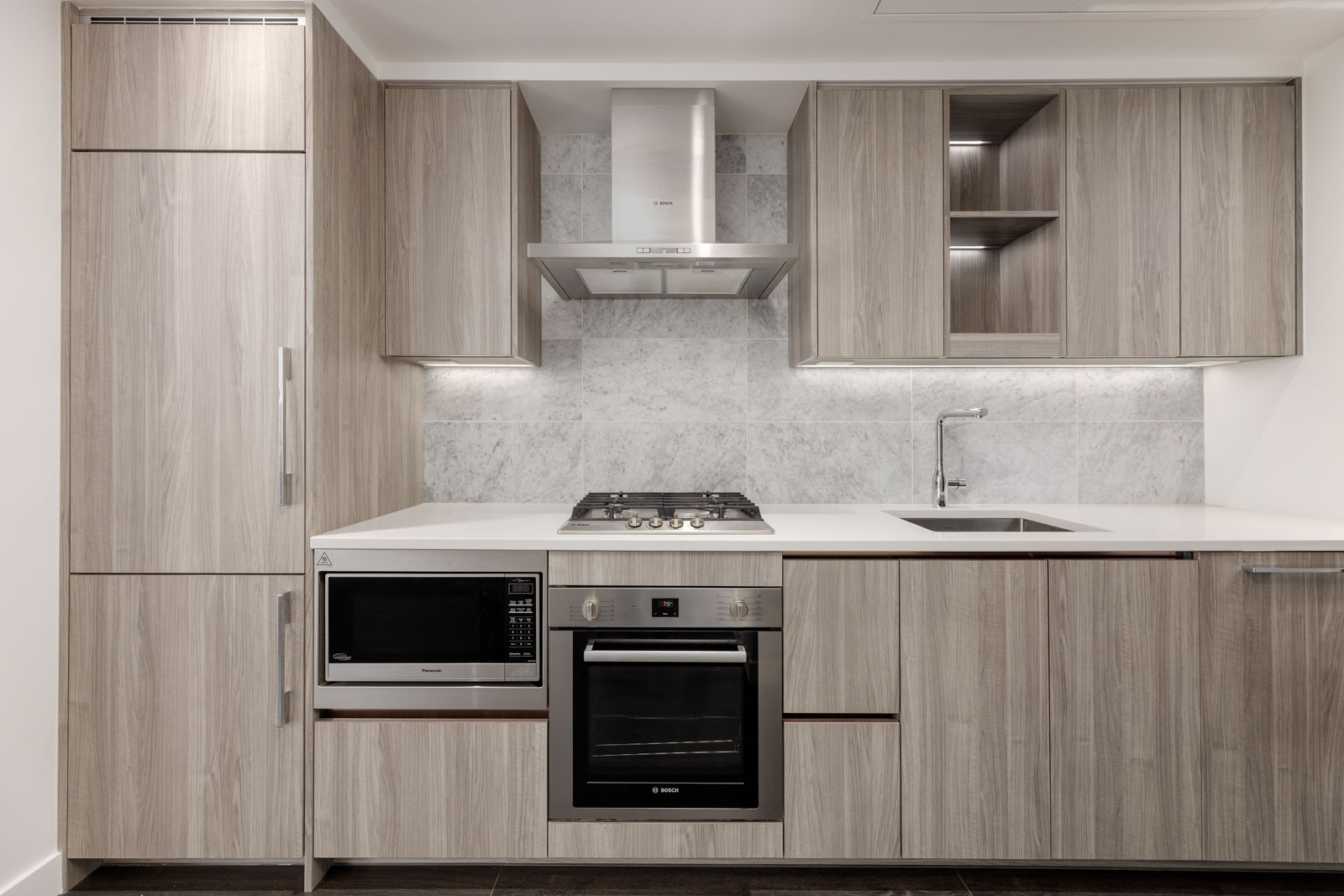 Modern kitchen with light wood cabinets, built-in appliances including oven, microwave, gas cooktop, and range hood, white countertop, and marble backsplash.