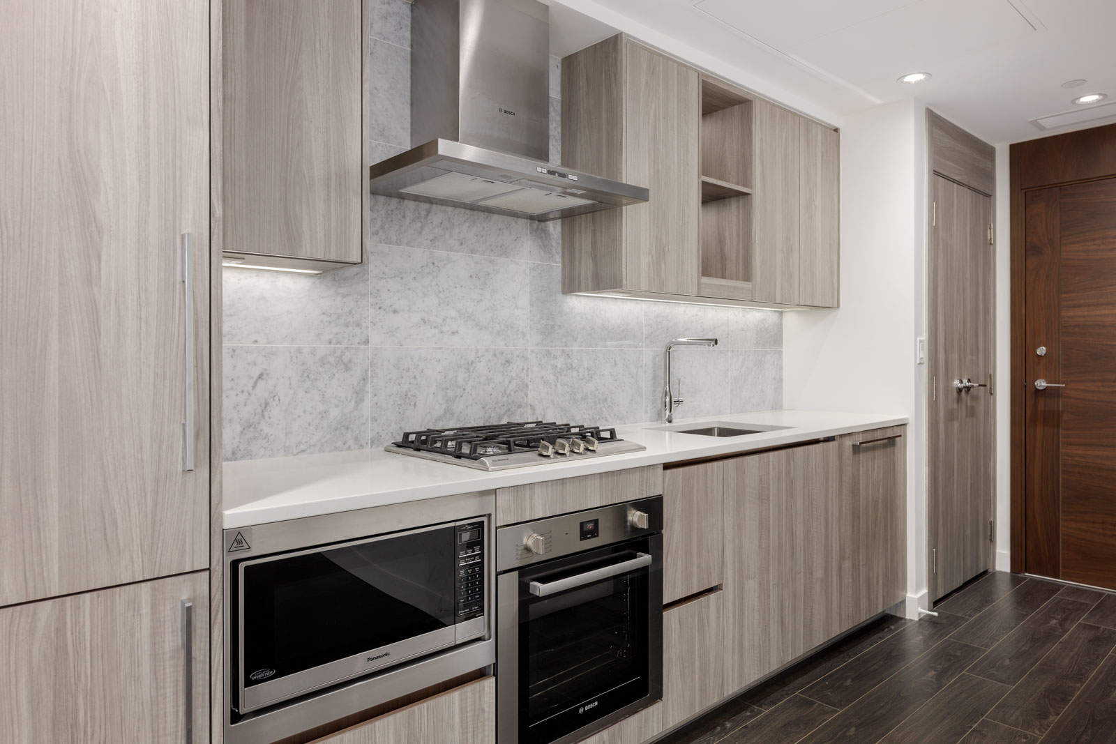 Modern kitchen with light wood cabinets, built-in microwave, oven, gas stove, stainless steel range hood, white countertop, and marble backsplash.